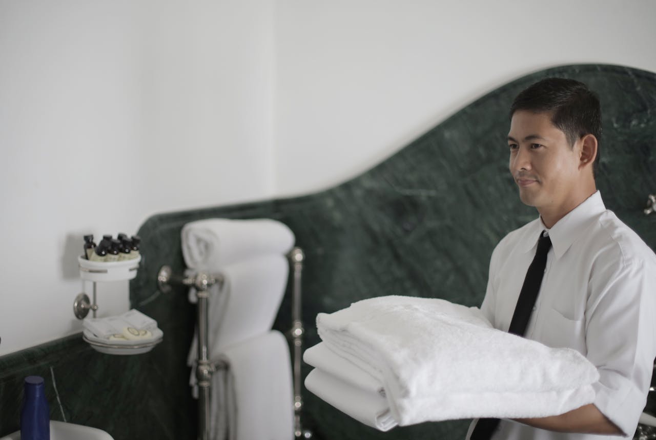 services-01 Side view of smiling ethnic male hotel employee holding stack of clean white towels while cleaning bathroom